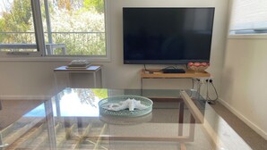 A modern living room with a glass coffee table, a flat-screen TV, and a view of trees through a large window. - 20 Bradley (Coles Bay)