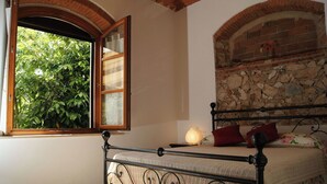 A bedroom with a bed, a window with an open shutter, a stone wall, and a lamp.