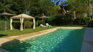 A swimming pool with clear water, surrounded by a well-maintained lawn and trees. There are lounge chairs and a gazebo with a canopy.