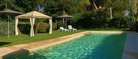 A swimming pool with clear water, surrounded by a well-maintained lawn and trees. There are lounge chairs and a gazebo with a canopy.