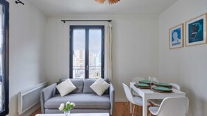 Una sala de estar moderna con un sofá gris, una mesa de comedor con sillas blancas y vistas de la ciudad a través de ventanas amplias.