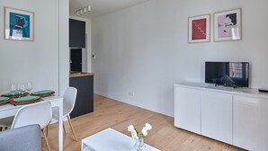 Una sala de estar moderna con comedor, una cocina al fondo y un televisor sobre un mueble blanco.