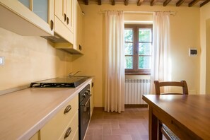 A kitchen with a stove, oven, and cabinets. There is a window with curtains, a radiator, and a table with a chair. - Beautiful farmhouse with swimming pool (Città di Castello)