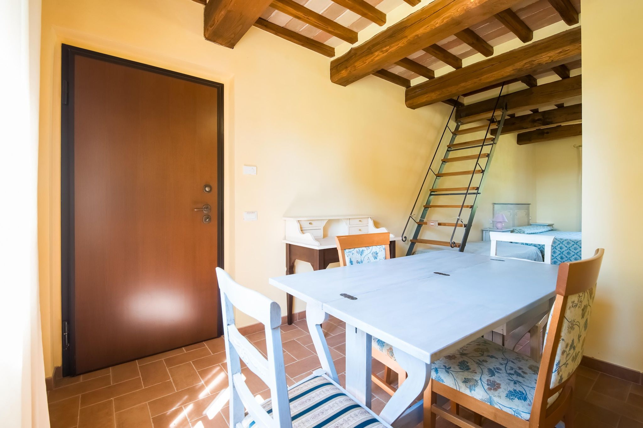Een kamer met een houten deur, een eettafel met stoelen, een klein bureau, een bed met een beddegoed met dessin, en een houten trap.