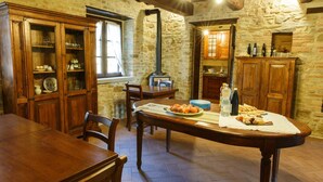 A rustic dining area with a wooden table, chairs, and a stone wall. There is a window providing natural light and a built-in wood stove.