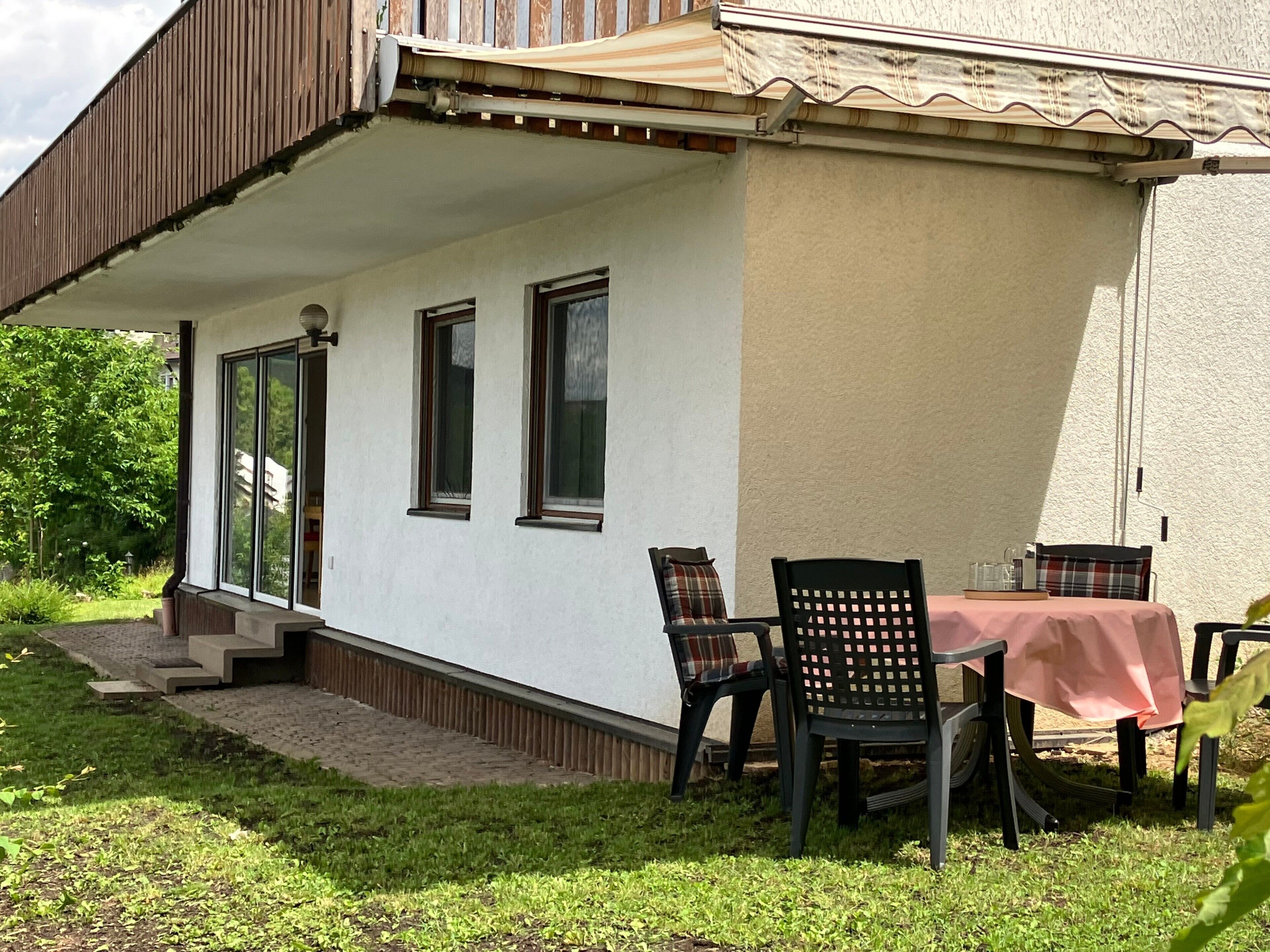 A covered outdoor seating area with a table and chairs, a small set of steps leading to a door, and a view of a garden.