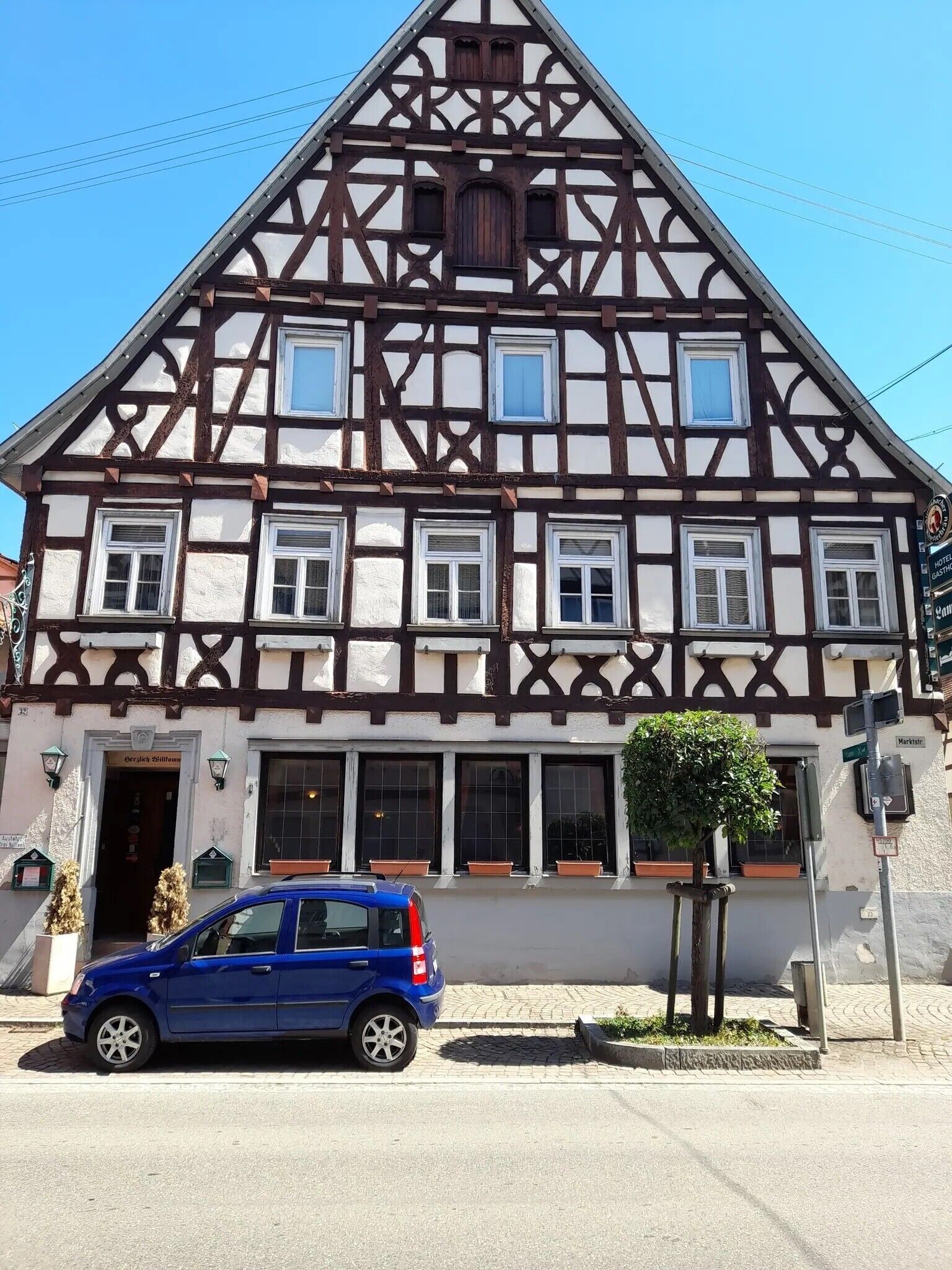 A traditional half-timbered building with multiple windows, a car parked in front, and a small tree in the foreground.