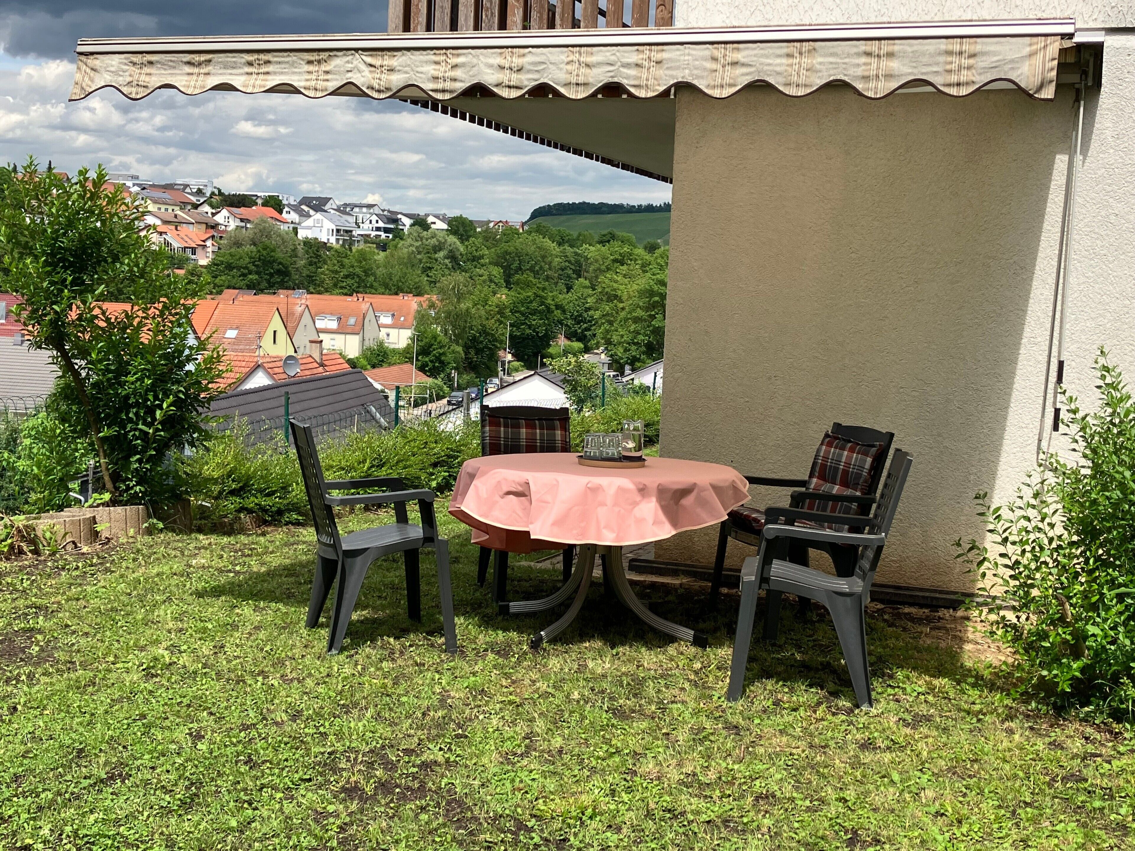 Eine Terrasse im Freien mit Tisch und Stühlen, umgeben von Grünpflanzen und mit Blick auf eine Wohngegend.
