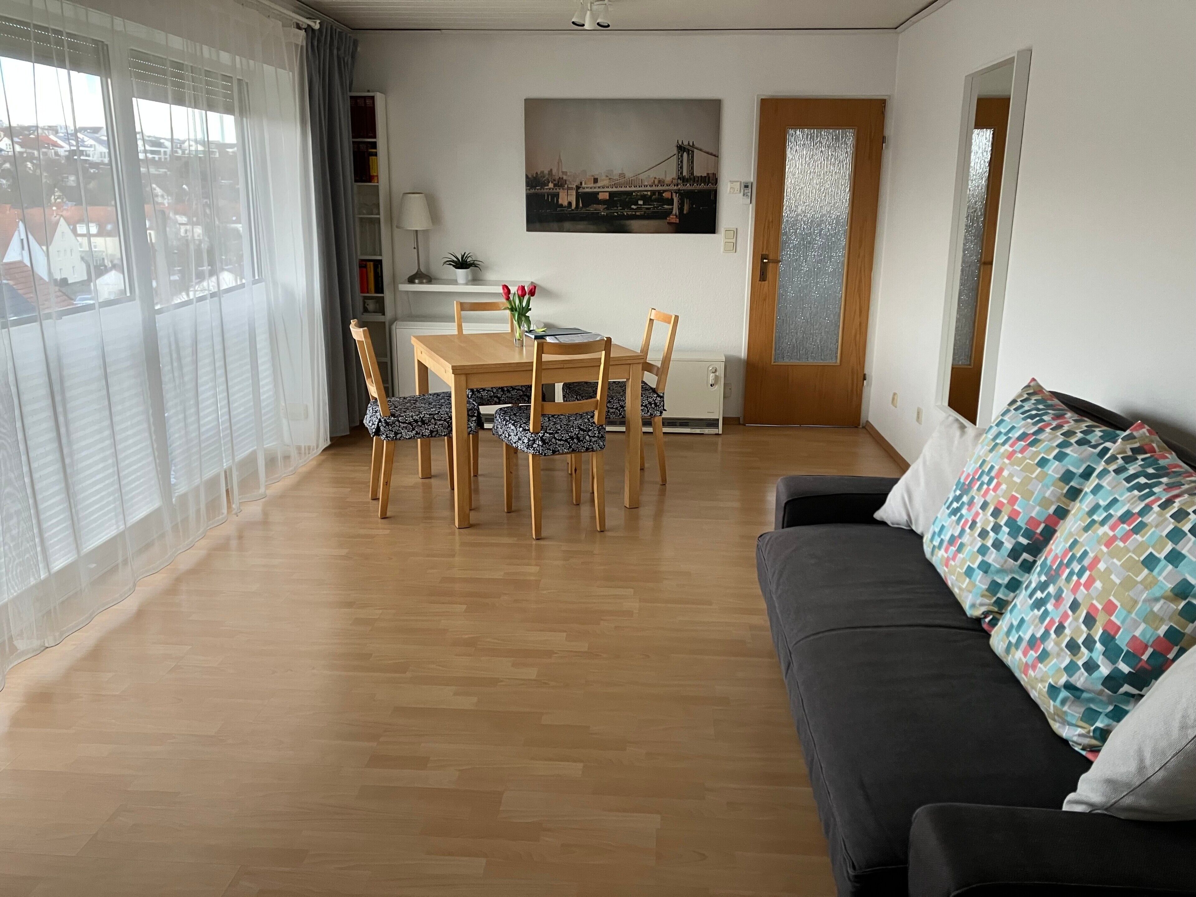 Ein Wohnzimmer mit Esstisch aus Holz, Stühlen, einer Couch, einer Topfpflanze und einem Stadtbild an der Wand.