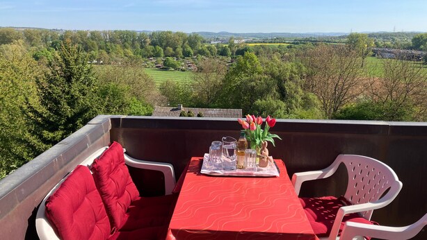 Un balcon avec une table dressée pour deux personnes, des coussins rouges et un vase rempli de fleurs.