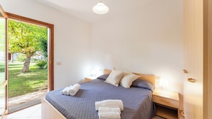 A bedroom with a bed, two bedside tables, a nightstand, and a view of a garden through a sliding door. - Cozy Apartment 300mt From the Beach (Budoni)