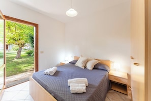 A bedroom with a bed, two bedside tables, a nightstand, and a view of a garden through a sliding door. - Cozy Apartment 300mt From the Beach (Budoni)