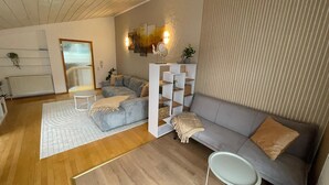 Moderni olohuone, jossa on harmaa sohva, valkoinen sohvapöytä ja valkoinen sivupöytä. Lattiassa on puulattia, valkoinen matto ja seinässä on raidallinen tapetti.