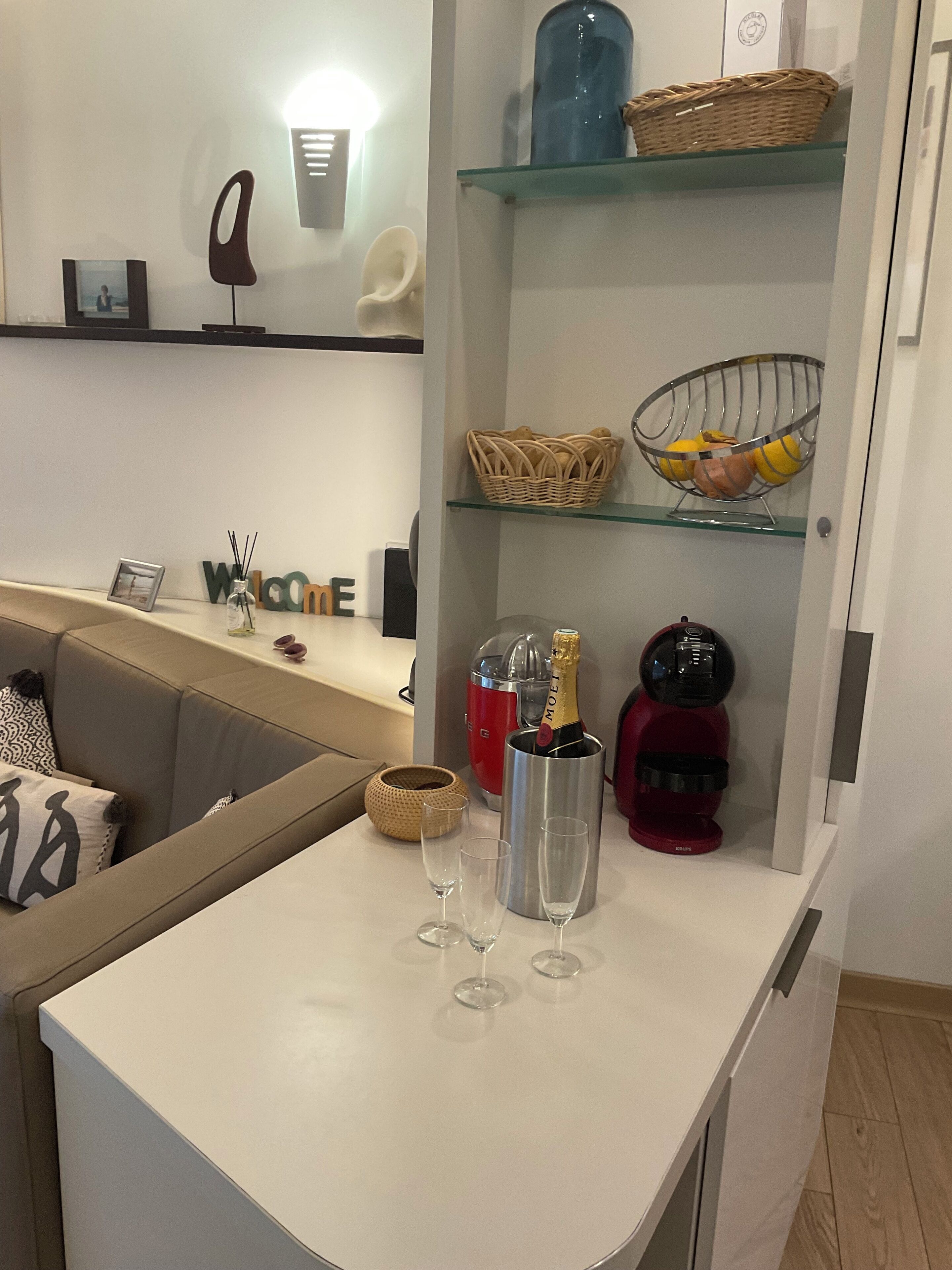 A modern kitchen with a white countertop, a wine cooler, a red coffee machine, and a glass shelf with baskets and a bowl.