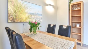 Ein Essbereich mit einem Holztisch, vier Stühlen, einem Blumenstrauß, einem Wandbild und einem Holzregal mit Gläsern.