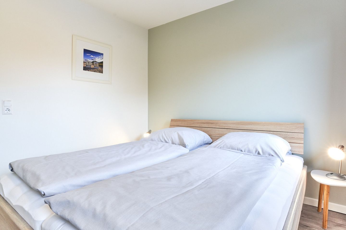 Ein Doppelbett mit Holz-Kopfteil, ein Nachttisch mit Lampe sowie ein gerahmtes Bild an der Wand.