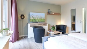 Hotelkamer met een bed, een bureau met een computer, een stoel, een bijzettafeltje met een bloevas en een groot raam met gordijnen.
