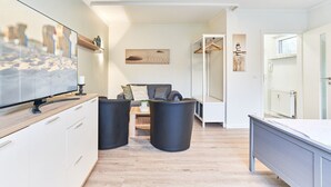 A modern, well-lit room with a flat-screen TV, a sofa, two black ottomans, a wooden table, and a coat rack. - Haus SeeZeit, Apartment 03 - Haus SeeZeit (Timmendorfer Strand)