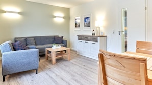 Un salon moderne avec un canapé gris, une table basse en bois et un coin repas comprenant une table et des chaises en bois. On y trouve également un meuble blanc avec des tiroirs et un petit plan de travail.