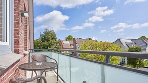 Een balkon met rieten meubilair, een glazen balustrade en uitzicht op omliggende huizen en bomen.