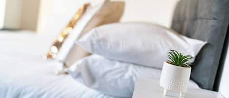 A neatly made bed with white linens and a grey headboard. There is a white bedside table with a small potted plant on it.