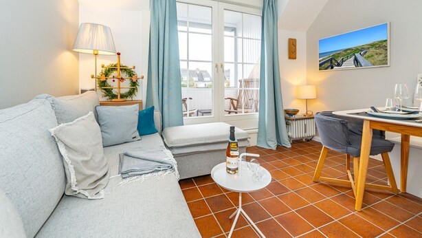 A cozy living room with a sofa, a dining table set for two, a window with curtains, and a painting on the wall. - Syltweh - Comfortable Syltweh vacation apartment with south-facing balcony in Munkmarsch (Sylt)
