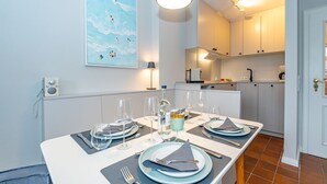 A dining area with a table set for two, a kitchen in the background, and a painting on the wall. - Syltweh - Comfortable Syltweh vacation apartment with south-facing balcony in Munkmarsch (Sylt)