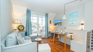 A modern living room with a sofa, dining table, and chairs. There is a window with curtains, a lamp, and a painting on the wall. - Syltweh - Comfortable Syltweh vacation apartment with south-facing balcony in Munkmarsch (Sylt)