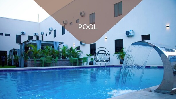A swimming pool with a water feature, surrounded by a building with air conditioning units and outdoor seating. - Jasmine Luxury Apartment (Ikorodu)