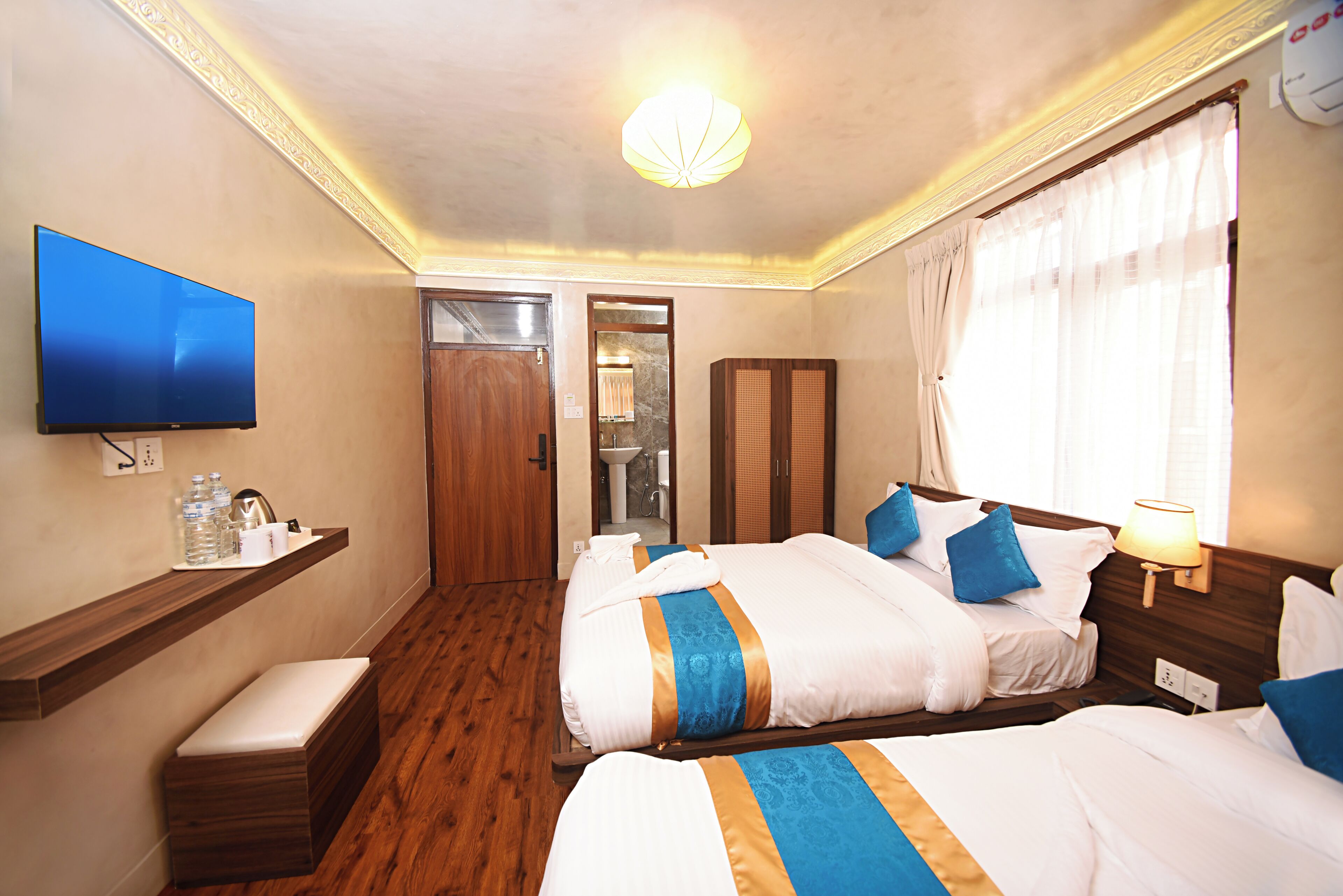 A hotel room with two beds, a TV, a wooden bench, a shelf with toiletries, and a bathroom visible through an open door.