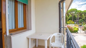 Ein Balkon mit weißem Tisch und Stuhl, ein Fenster mit Holzläden und Blick auf eine Straße mit Bäumen und einer Fußgängerampel.