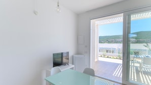 Ein modernes Zimmer mit Glastisch, weißen Stühlen, einem Flachbildfernseher und Blick auf einen Balkon mit Gartenmöbeln.