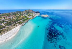 An aerial view of a coastal area with a beach, clear turquoise waters, and a rocky outcrop.