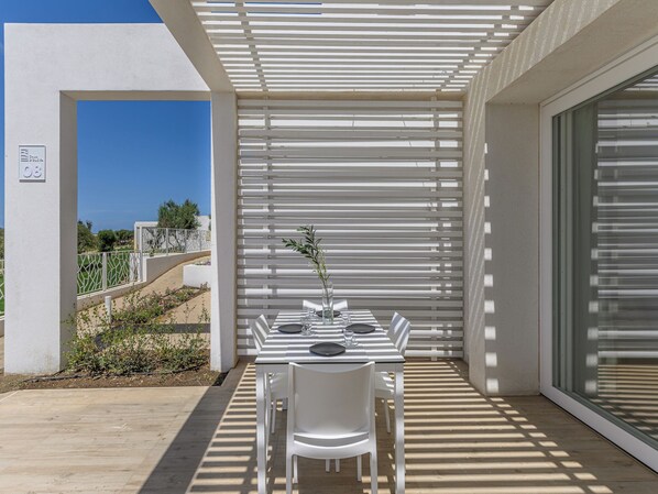 An outdoor patio with a white table set for two, surrounded by white chairs, under a white pergola.