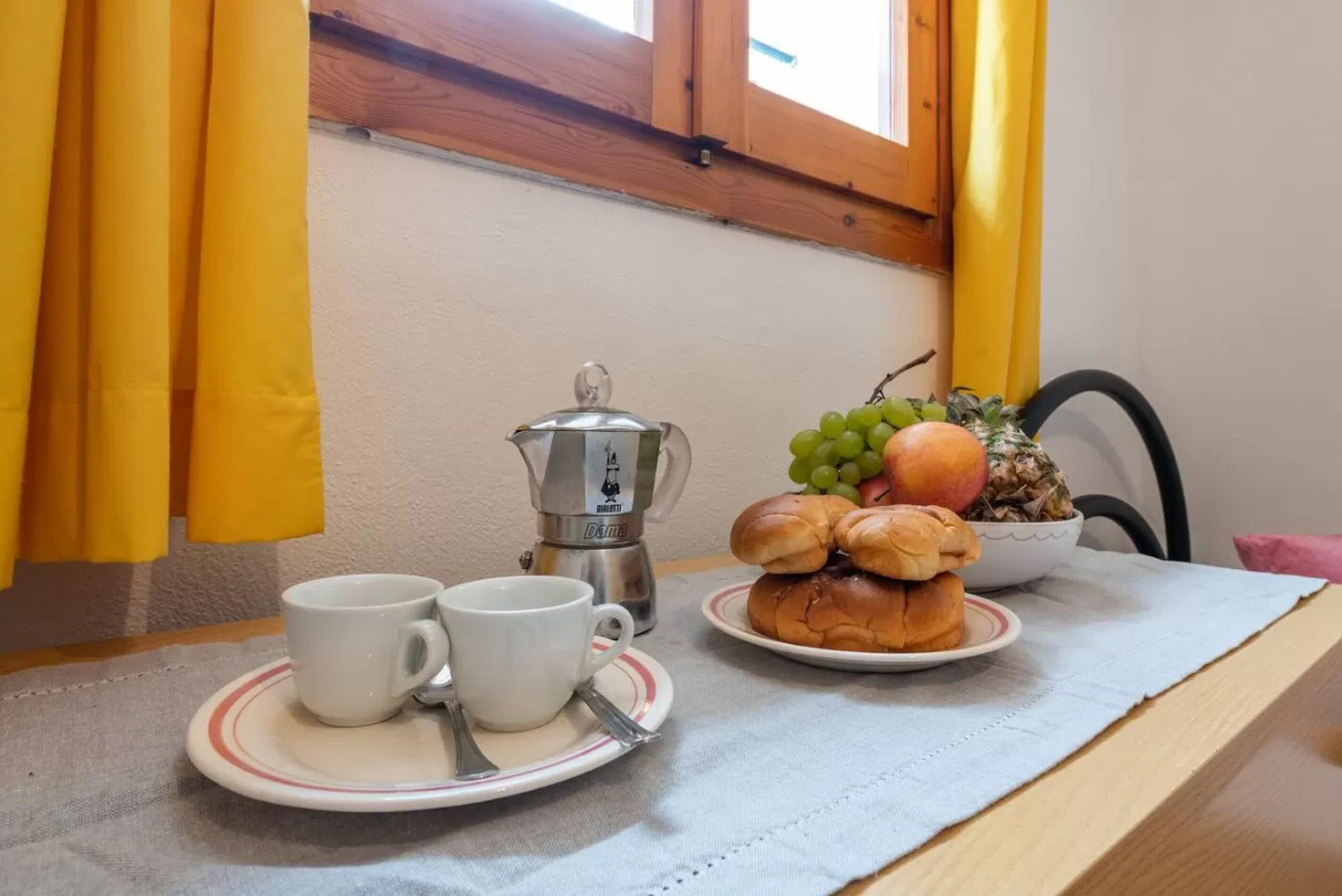 En morgenmadsanretning med en kaffemaskine, to kopper, en tallerken med brød og frugt, samt et vindue med gardiner.