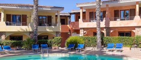 A pool area with lounge chairs and a clear blue pool, surrounded by multi-story buildings with balconies and windows.