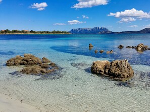 A clear coastal beach with visible rocks and a distant mountain range. - Cozy Apartment 150 m From Pittulongu Beach (Olbia)