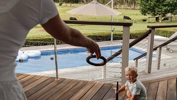 A wooden deck with a pool and a grassy area with trees and a fence in the background.