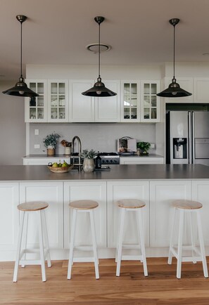 A modern kitchen with white cabinetry, a stainless steel refrigerator, and three bar stools. - Riverglen Country Escape Pool and River Views Sleeps 10 (Glenthorne)