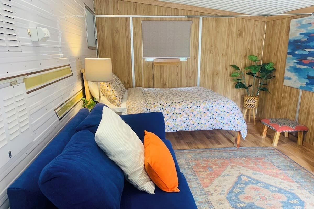 A compact bedroom with a blue sofa, a bed with a floral bedspread, a wooden wall panel, a small plant, and a colorful rug.
