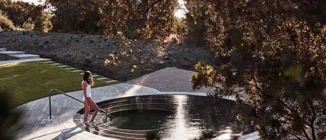 A person standing by a circular water feature in a landscaped area with a paved walkway and a grassy lawn.