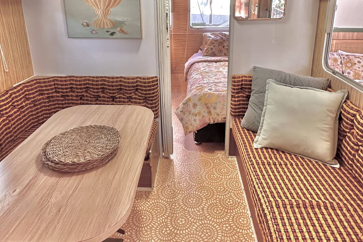 A compact RV interior with a dining area, a bed with patterned bedding, and a wall-mounted artwork.