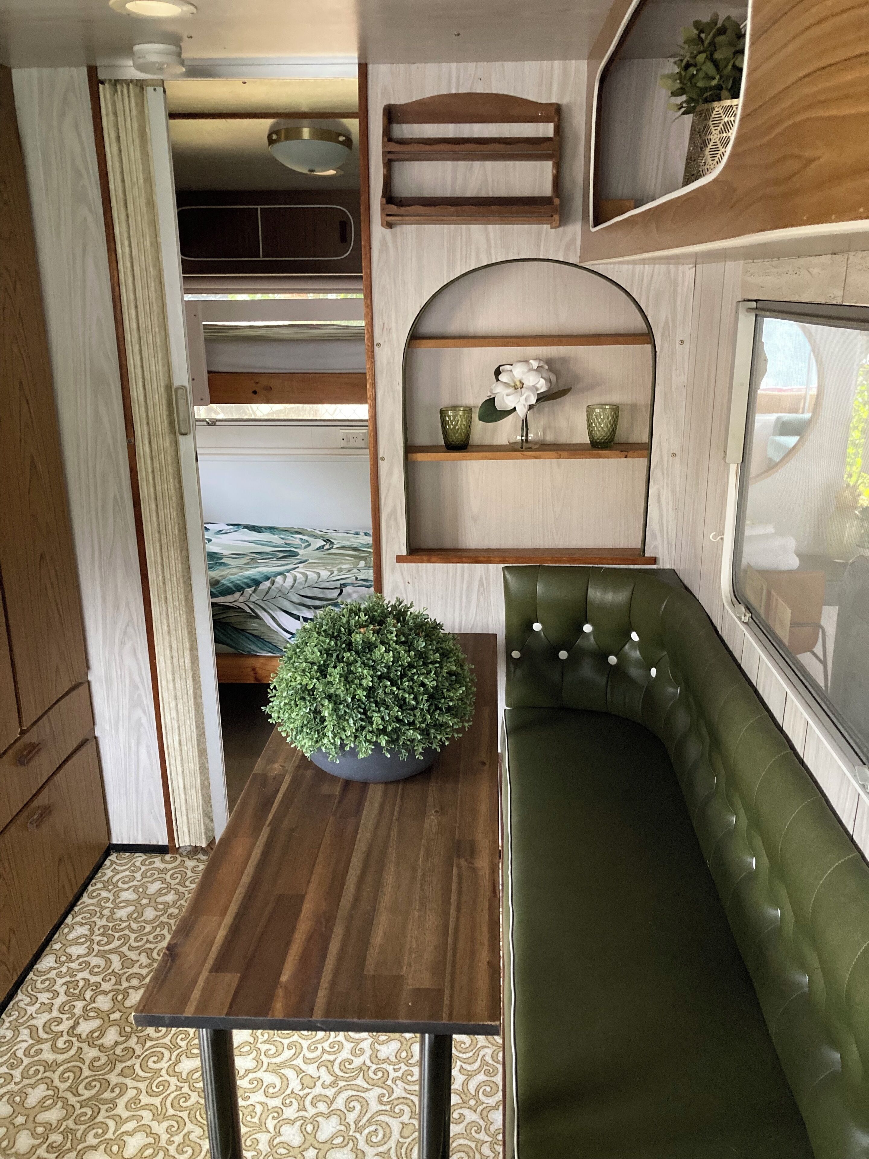 A compact, well-organized interior with a bunk bed, a green cushioned seating area, a small table with a potted plant, and built-in shelves with decorative items.