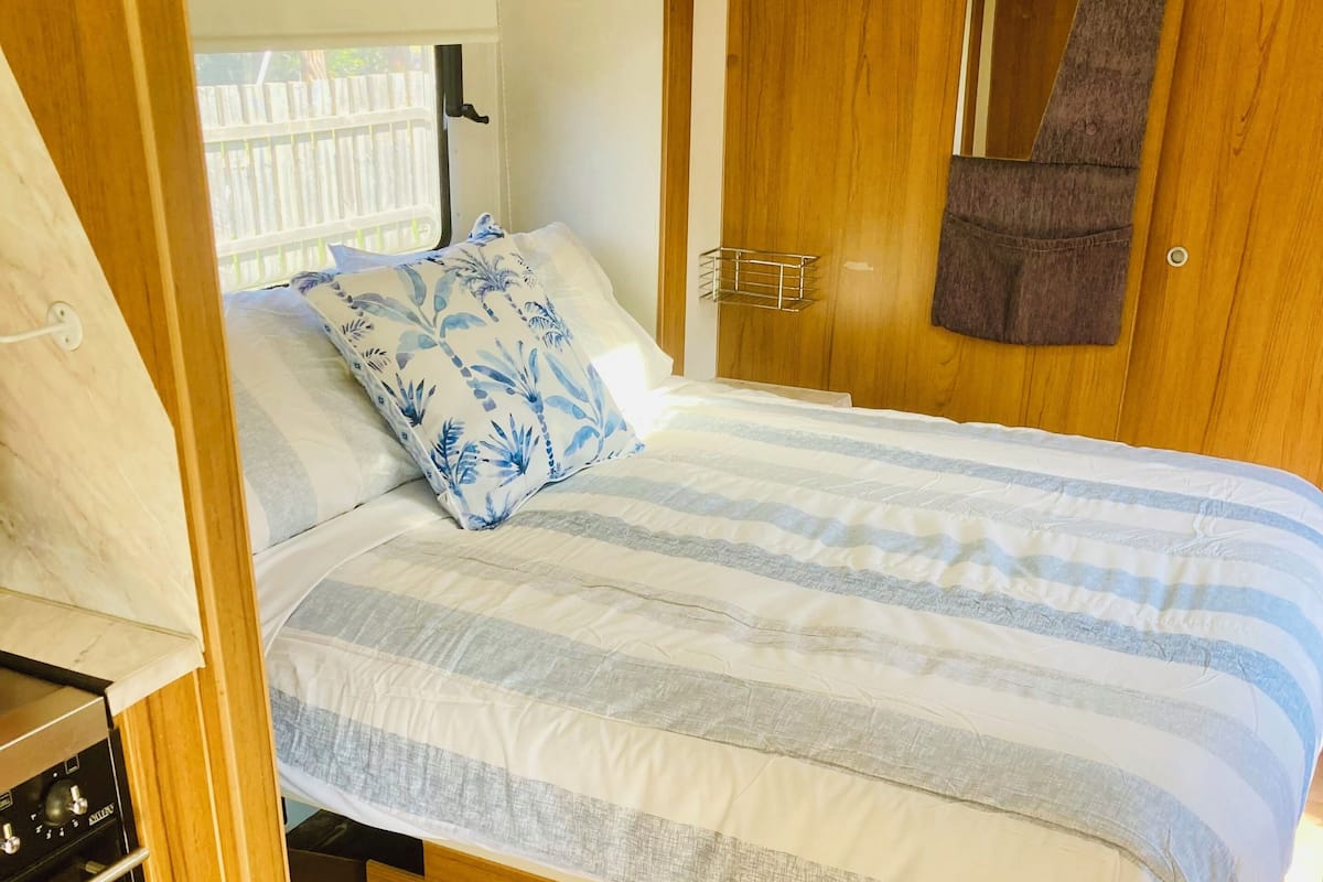 A compact, wood-paneled bedroom with a bed, a small kitchenette, and a window with a view of a fence.