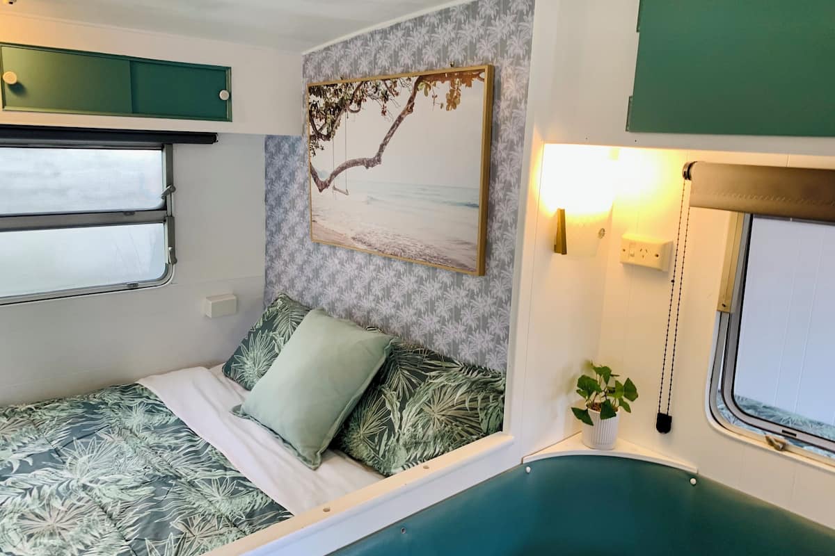 A compact, well-lit room with a bed featuring a leaf-patterned bedspread, a wall-mounted painting of a tree branch, a small potted plant, and a window with a view of the outside.
