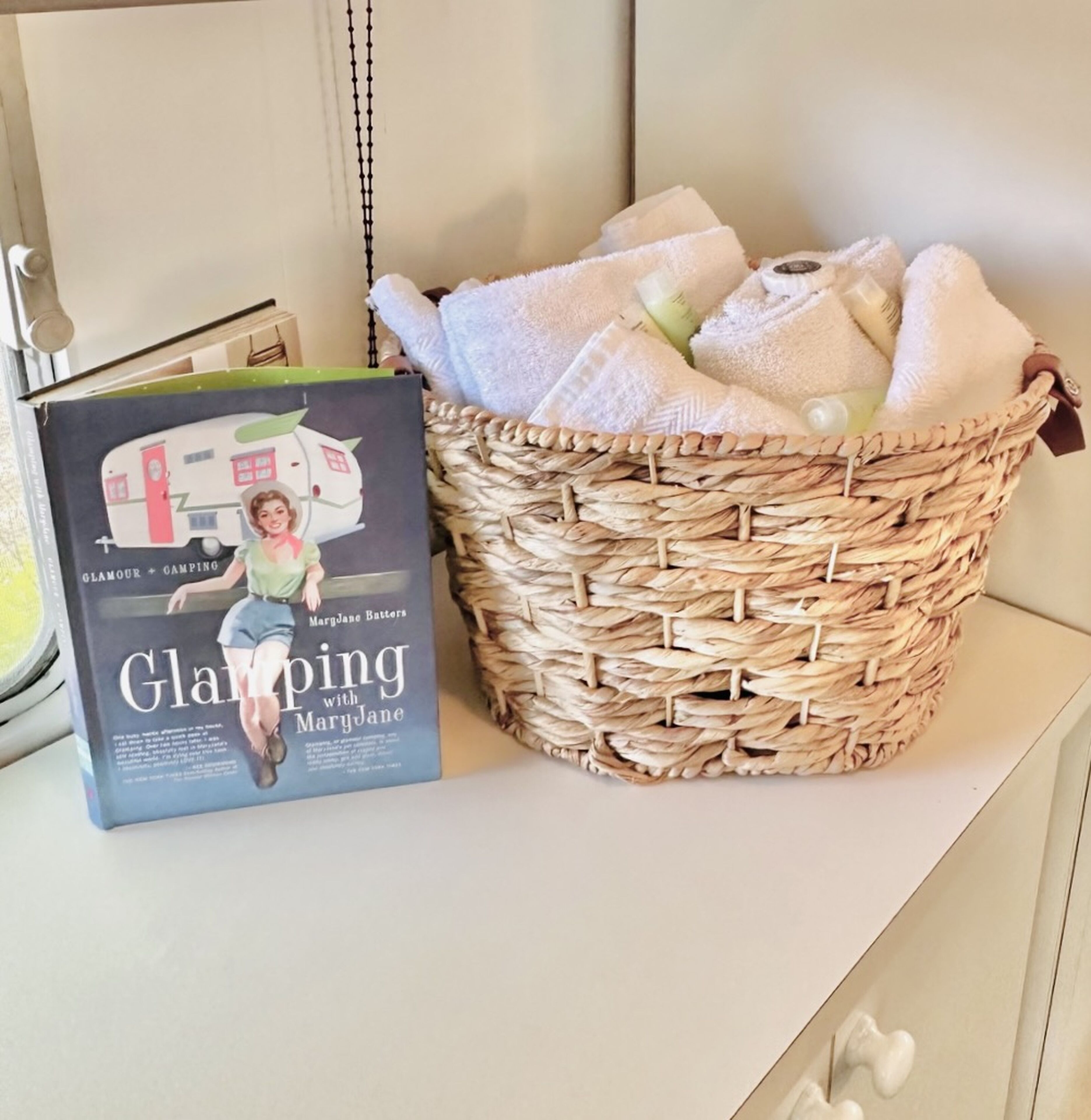 A wicker basket filled with white towels and a book titled 'Glamping with MaryJane' on a shelf.