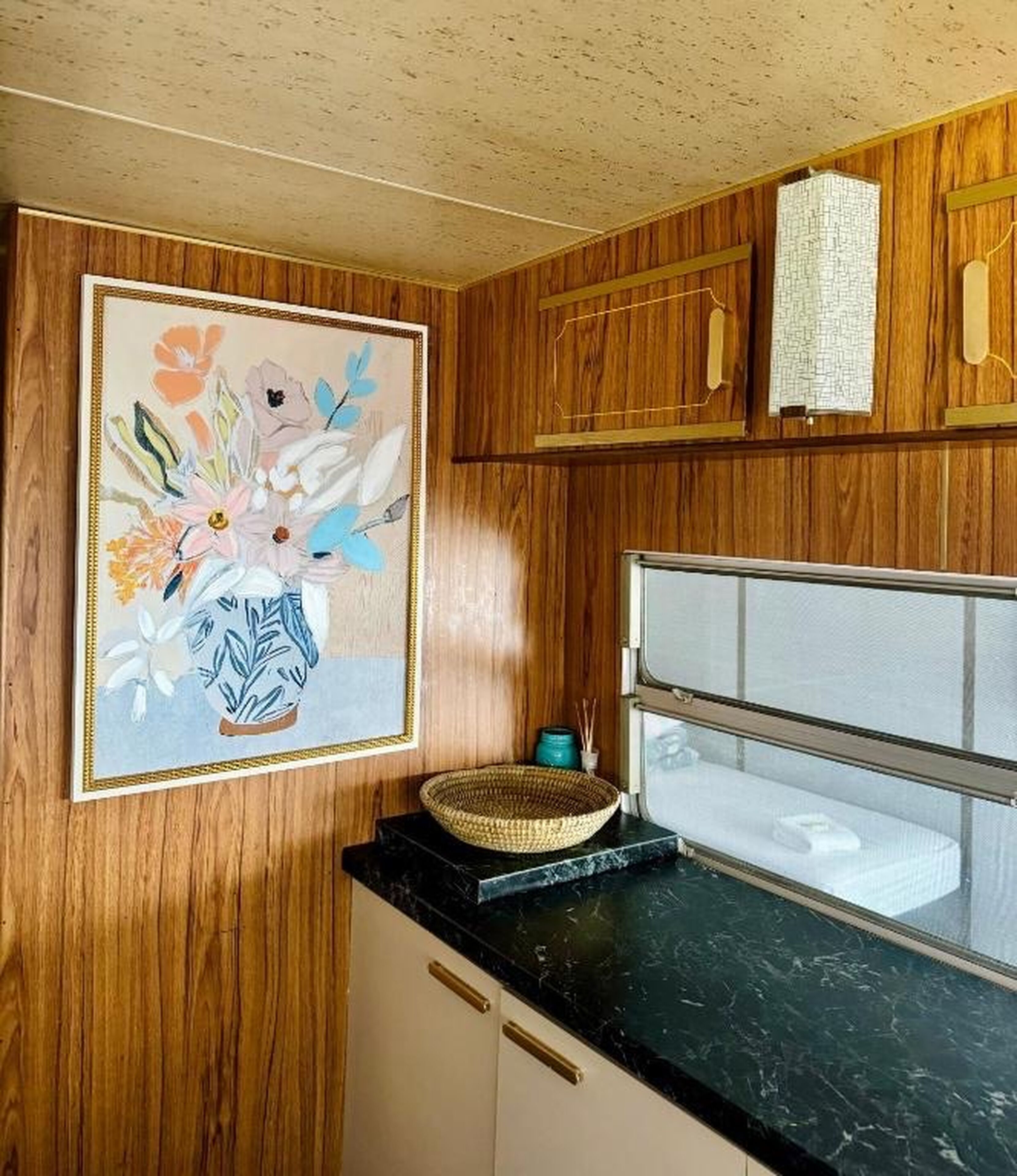 A kitchen with wooden cabinets, a marble countertop, a framed floral artwork, and a basket.