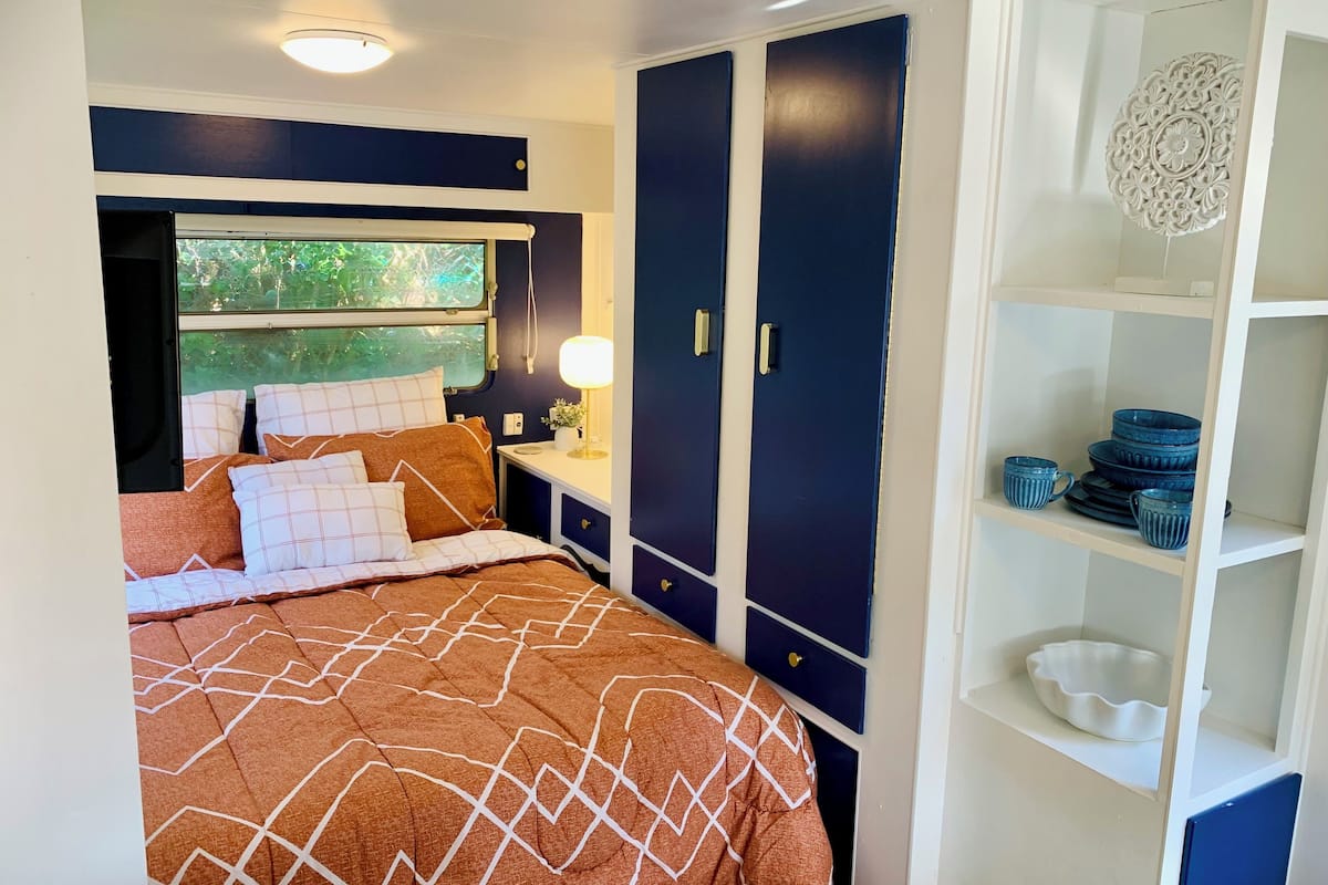 A bedroom with a bed, a nightstand, a lamp, a wardrobe, and a shelf with dishes.