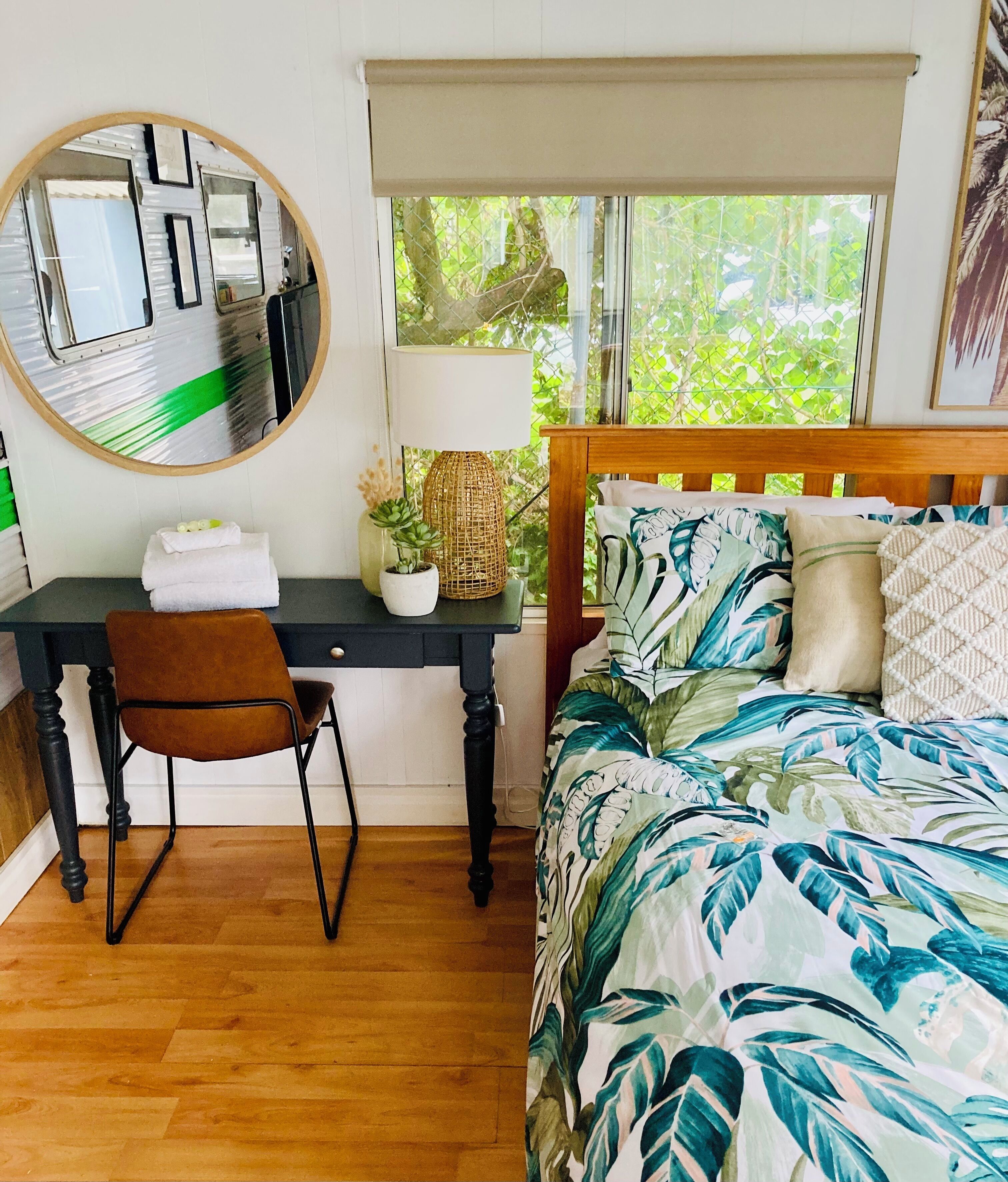 A bedroom with a bed, a desk, a chair, a mirror, and a window with a view of greenery.