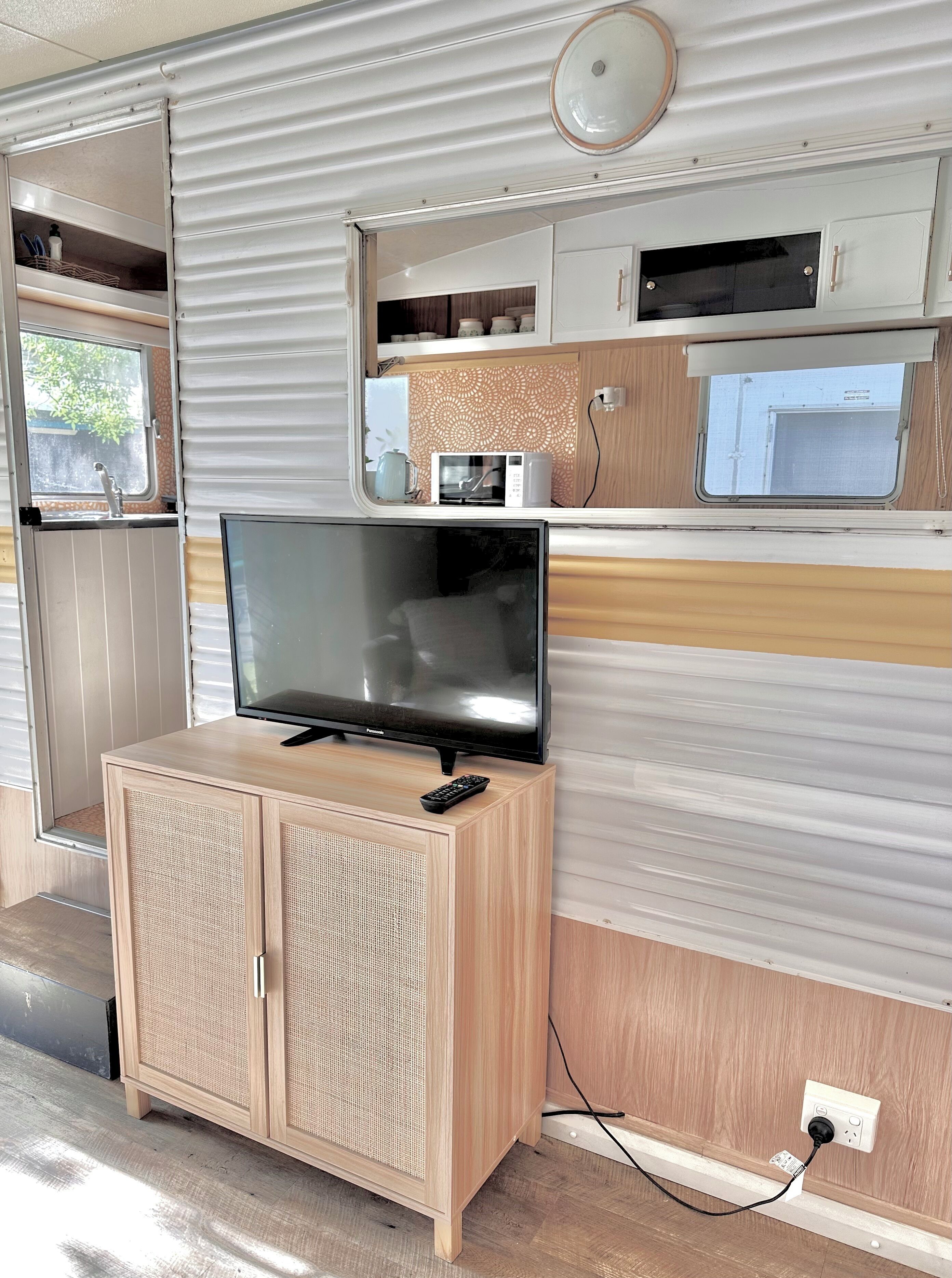 Een compact interieur van een camper met een televisie op een houten kast, een magnetron op een plank, en een raam met uitzicht op de omgeving.
